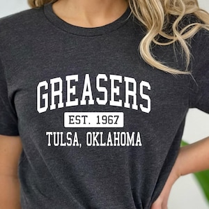 May include: Dark gray t-shirt with the word "GREASERS" in a white, arched font. Below, the text reads "EST. 1967" and "TULSA, OKLAHOMA" in a white, block font. The shirt is a crew neck style.
