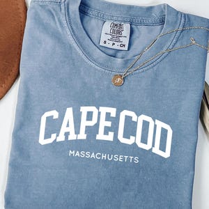 May include: A blue t-shirt with the text "CAPE COD MASSACHUSETTS" in white block letters. The shirt has a crew neck and short sleeves. A gold necklace with a pendant is draped on the shirt.