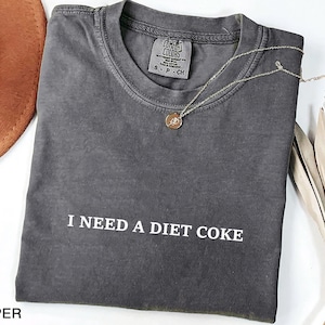 May include: A dark gray t-shirt with the text "I NEED A DIET COKE" in white. The shirt is folded and has a small tag at the neckline. A gold necklace with a round pendant rests on the shirt. A brown hat and dried plant are in the background.