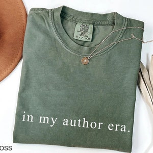 May include: An olive green t-shirt with the text "in my author era." A gold necklace with a pendant rests on the shirt. The shirt is labeled "Comfort Colors" and is size small. A brown hat and dried plant are in the background.