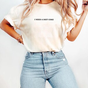 May include: Cream-colored t-shirt with the text "I NEED A DIET COKE" in black. The shirt is paired with light blue denim jeans. The overall style is casual and relaxed.