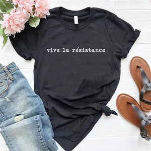 May include: Black t-shirt with the phrase "vive la r&eacute;sistance" in white lettering. The shirt is paired with blue jeans, brown sandals, and pink flowers. A casual, everyday outfit.
