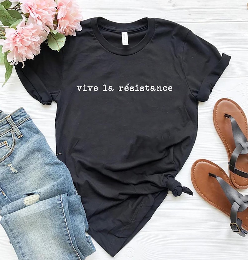 May include: Black t-shirt with the phrase "vive la r&eacute;sistance" in white lettering. The shirt is paired with blue jeans, brown sandals, and pink flowers. A casual, everyday outfit.