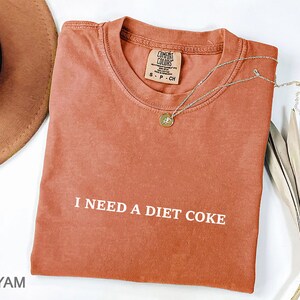 May include: A rust-colored t-shirt with the phrase "I NEED A DIET COKE" printed in white. A gold-colored necklace with a pendant rests on the shirt. The shirt is folded and has a tag that reads "COMFORT COLORS".