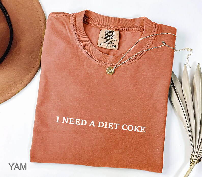 May include: A rust-colored t-shirt with the phrase "I NEED A DIET COKE" printed in white. A gold-colored necklace with a pendant rests on the shirt. The shirt is folded and has a tag that reads "COMFORT COLORS".