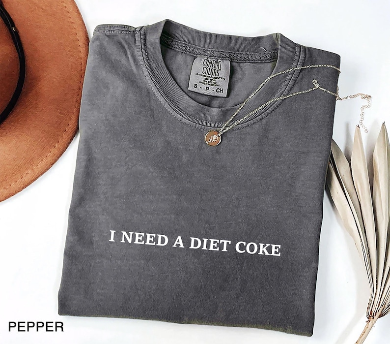May include: A dark gray t-shirt with the text "I NEED A DIET COKE" in white. The shirt is folded and has a small tag at the neckline. A gold necklace with a round pendant rests on the shirt. A brown hat and dried plant are in the background.