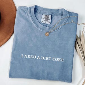 May include: A blue t-shirt with the text "I NEED A DIET COKE" in white. A gold necklace with a pendant rests on the shirt. The shirt is folded, with a brown hat and dried plant in the background.