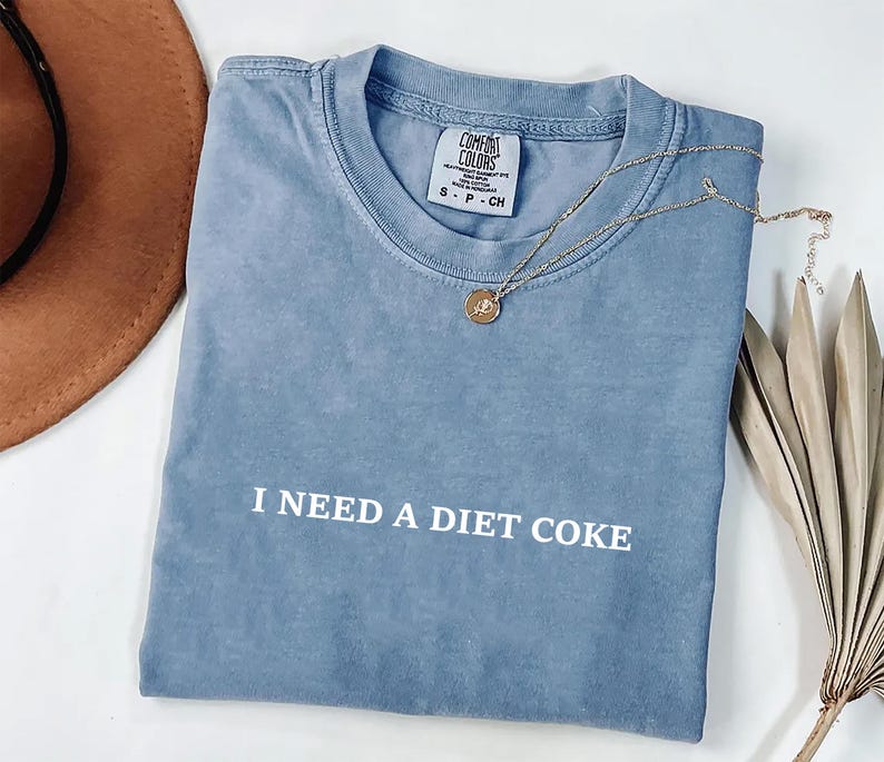 May include: A blue t-shirt with the text "I NEED A DIET COKE" in white. A gold necklace with a pendant rests on the shirt. The shirt is folded, with a brown hat and dried plant in the background.