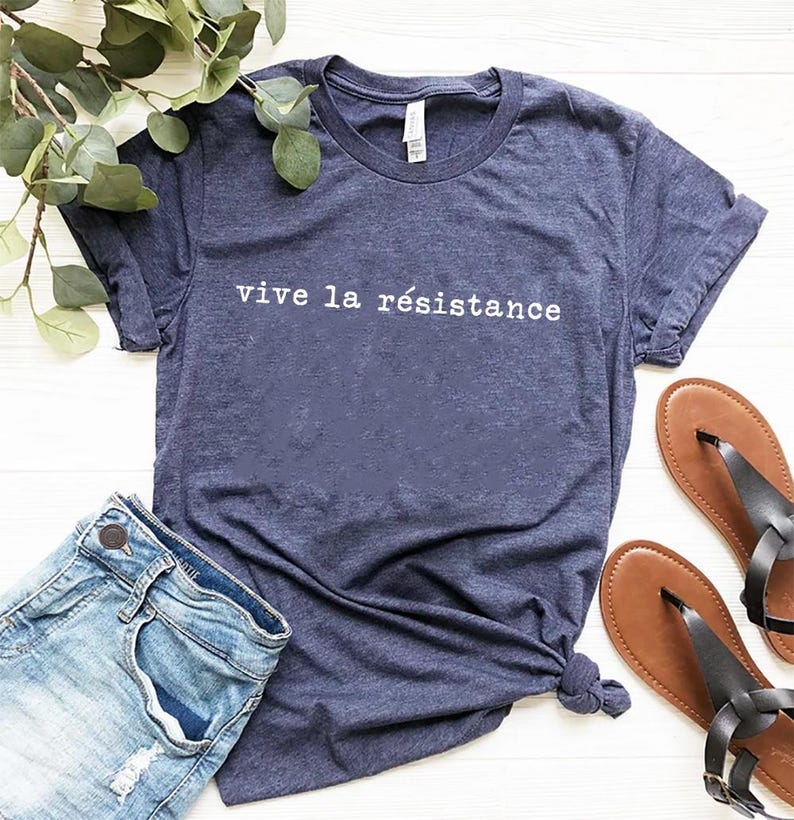 May include: A navy blue t-shirt with the phrase "vive la r&eacute;sistance" printed in white. The shirt is paired with denim shorts and brown sandals. The items are arranged on a white surface with green foliage.