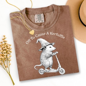 May include: Brown t-shirt with the text "Off To Cause A Kerfuffle" and a graphic of an opossum wearing a witch hat and riding a scooter. A gold heart pendant necklace and a tan hat are also visible.