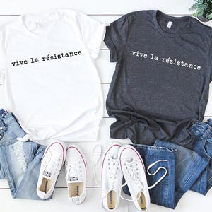 May include: Two t-shirts, one white and one charcoal gray, each with the phrase "vive la r&eacute;sistance" printed in black. The shirts are paired with blue jeans and white sneakers with red accents.