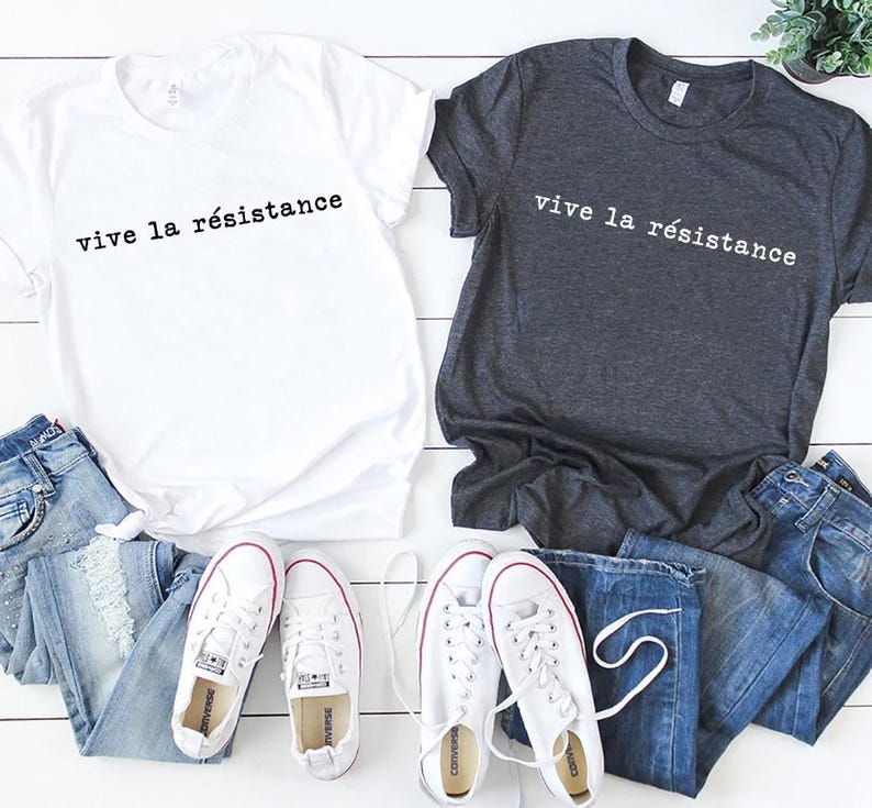 May include: Two t-shirts, one white and one charcoal gray, each with the phrase "vive la r&eacute;sistance" printed in black. The shirts are paired with blue jeans and white sneakers with red accents.