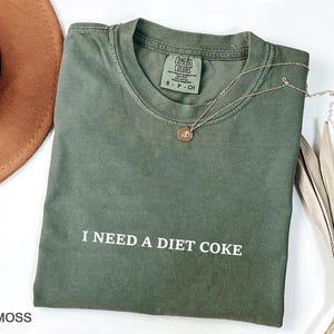 May include: Moss green t-shirt with the text "I NEED A DIET COKE" in white. A gold necklace with a round pendant rests on the shirt. The shirt is folded and has a Comfort Colors tag.