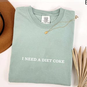 May include: A light green t-shirt with the text "I NEED A DIET COKE" in white. A gold necklace with a pendant rests on the shirt. A brown hat and dried palm leaves are in the background.