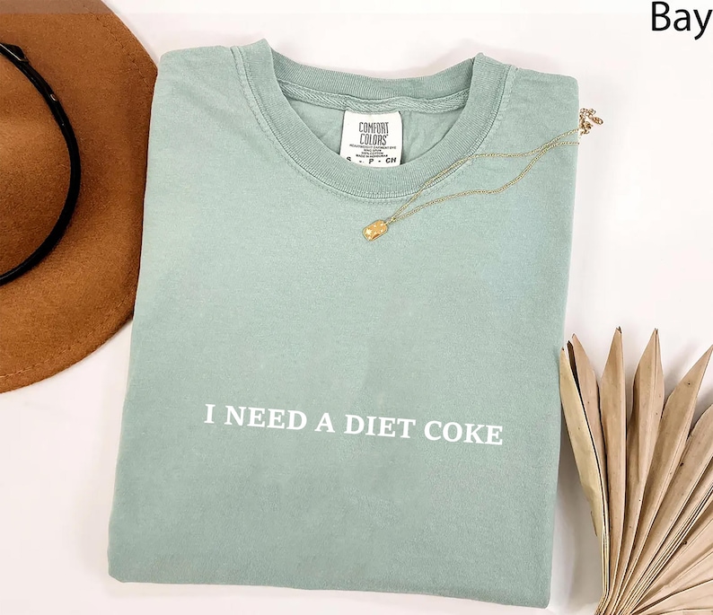 May include: A light green t-shirt with the text "I NEED A DIET COKE" in white. A gold necklace with a pendant rests on the shirt. A brown hat and dried palm leaves are in the background.