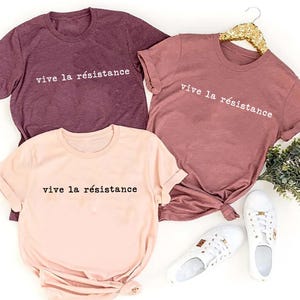 May include: Three t-shirts in shades of mauve, rose, and peach, each bearing the phrase "vive la r&eacute;sistance" in white lettering. White sneakers are placed near the peach shirt. The shirts are displayed on a white surface.