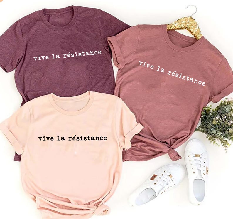 May include: Three t-shirts in shades of mauve, rose, and peach, each bearing the phrase "vive la r&eacute;sistance" in white lettering. White sneakers are placed near the peach shirt. The shirts are displayed on a white surface.