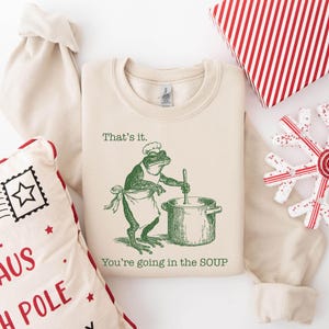 May include: Cream-colored sweatshirt with a green frog chef illustration stirring a pot, and the text "That's it. You're going in the SOUP." The image includes a gift box, a pillow, and a snowflake decoration.