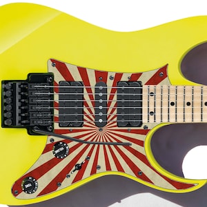 Può includere: Chitarra elettrica gialla con un design a raggiera rosso e bianco sul corpo. La chitarra ha un battipenna nero e una tastiera in acero.