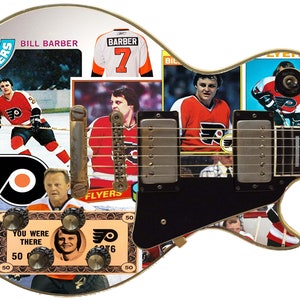 Peut inclure: Une guitare électrique blanche avec un pickguard noir et un collage de joueurs et de logos des Flyers de Philadelphie. La guitare est décorée d'images de joueurs en uniforme, du logo de l'équipe et des mots "Flyers" et "Bill Barber".