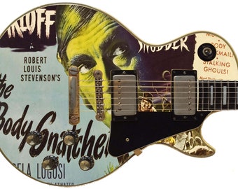May include: A black electric guitar with a custom design featuring a vintage movie poster for "The Body Snatcher" starring Bela Lugosi. The poster is in shades of blue, green, and yellow. The guitar has a black pickguard and a silver bridge.