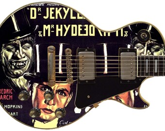 May include: A black electric guitar with a custom design featuring a vintage movie poster for "Dr. Jekyll and Mr. Hyde". The poster features two men in top hats, one with a sinister grin and the other with a more neutral expression. The guitar has gold hardware and a black pickguard.