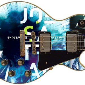May include: A black electric guitar with a blue, white, and green abstract design. The guitar has the text "Joe Satriani" and "Shockwave Supernova" on the body.