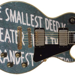 Könnte beinhalten: Eine blaue E-Gitarre mit einer weißen Aufschrift, die "Die kleinste Tat ist größer als die größte" lautet, mit goldenen Beschlägen und einem schwarzen Schlagbrett.