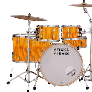 Puede incluir: Un set de batería con un acabado de madera amarilla. El bombo tiene el texto "Sticka Steves" impreso en él. El set incluye un bombo, un tambor de caja, dos toms y un hi-hat.