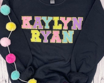 May include: Black sweatshirt with the names "KAYLYN RYAN" in colorful block letters with gold glitter accents. The sweatshirt is decorated with a garland of colorful pom-poms.