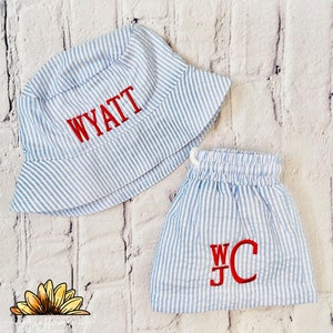 May include: A blue and white striped bucket hat with the name Wyatt embroidered in red. A matching blue and white striped pair of shorts with the initials WC embroidered in red.
