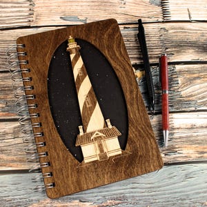 Op de afbeelding: Een bruin houten spiraalgebonden notitieboek met een lasergesneden ontwerp van een vuurtoren met zwart-witte strepen tegen een zwarte achtergrond met witte sterren.