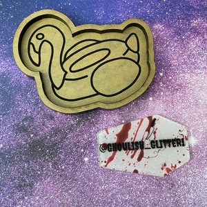 May include: A wooden mold shaped like a flamingo pool float. The mold is brown and has a black outline. A white resin piece with red blood splatter and a knife graphic is in the background. The resin piece has the text "@ghoulish_glitter1" on it.