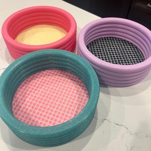May include: Three colorful, round trays with textured sides. One is pink with a yellow interior, another is teal with a pink patterned interior, and the third is purple with a black and white patterned interior. The trays are likely for home decor or organization.