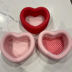 May include: Three heart-shaped containers in shades of red and pink. The red container is at the top, while two pink containers are below. The interiors have a woven pattern, with one pink container having a red and white checkered pattern.