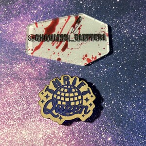 May include: Two decorative pins. The top pin is a clear, coffin-shaped acrylic with red splatter and the text "@ghoulish_glitter". The bottom pin is a wooden pin with the text "Thrift Queen" and a globe design in blue.