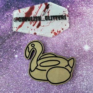 May include: A brown wooden cutout of a flamingo pool float. The cutout is on a purple glittery background. A clear plastic coffin with red blood splatter and a knife is in the background. The coffin has the text "@ghoulish_glitter1" on it.
