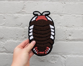 May include: A brown, white, red, and black embroidered patch in the shape of a pill bug. The patch has a textured appearance with a detailed design. The pill bug has a segmented body and is held against a white brick wall.