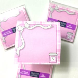 May include: Pink 3x3 inch Post-it notes with a white bow and ribbon design. The notes have a pink and white gingham border and a small Rx symbol. Each pad contains 50 sheets.
