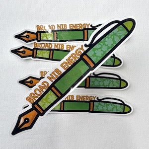 May include: A stack of die-cut stickers shaped like fountain pens. Each sticker features a green barrel with a floral pattern, a gold-colored nib, and the text BROAD NIB ENERGY printed in orange letters along the side of the pen body.
