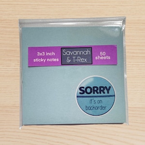 May include: A package of 3x3 inch sticky notes with the text "Savannah & T-Rex" and "50 sheets" on a light blue background. A circular sticker reads "SORRY it's on backorder". The sticky notes are in a clear plastic sleeve.