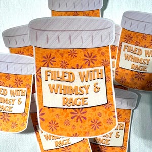 May include: A collection of stickers shaped like prescription pill bottles. The bottles are orange with a floral pattern and the text "FILLED WITH WHIMSY & RAGE" on a white label. The lids are white and gray.