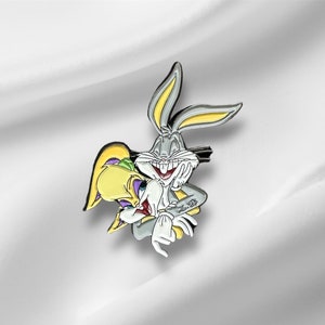 Może przedstawiać: Srebrna i żółta emaliowana przypinka przedstawiająca Bugs Bunny'a i Lolę Bunny'a, którzy się obejmują. Przypinka jest kreskówkową ilustracją postaci.