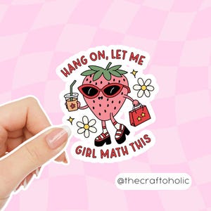 May include: A sticker featuring a cartoon strawberry wearing sunglasses and heels, holding a drink and a shopping bag. Text reads "Hang on, let me girl math this." The sticker is held by a hand against a pink checkered background.