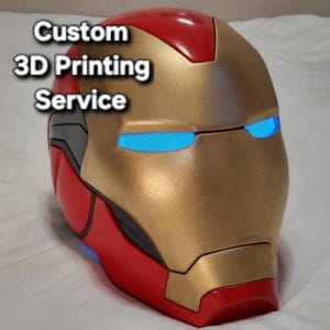 May include: A red and gold Iron Man helmet featuring glowing blue eyes and the text Custom 3D Printing Service. The helmet has a smooth, glossy finish with metallic gold faceplate detailing and dark grey accents along the side panels.