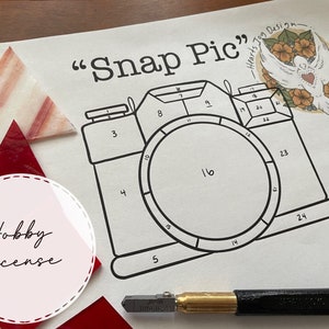 May include: A black and white line drawing of a camera with the words "Snap Pic" and "Hearts to Design" written above it. The camera is divided into 24 numbered sections. The image also includes the words "Hobby License" in a pink circle.