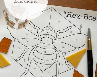 Puede incluir: Un dibujo en blanco y negro de una abeja con secciones numeradas en una hoja de papel blanca. La abeja está rodeada por una forma hexagonal. Hay piezas de vidrio naranja y amarillo en el papel, junto con una herramienta negra y dorada.