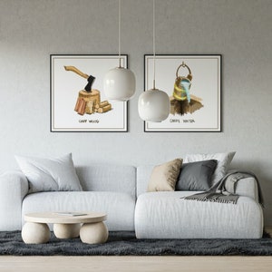 Peut inclure: Deux illustrations aquarelle encadrées accrochées à un mur au-dessus d'un canapé blanc. Les illustrations représentent un bloc de bois à hacher avec une hache et un seau d'eau. Le texte sous les illustrations indique "Camp Wood" et "Camp Water".