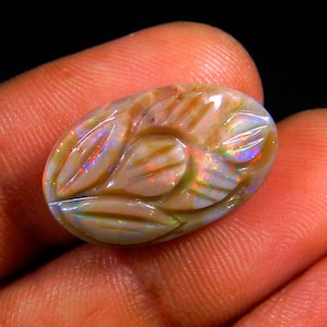 May include: An oval-shaped, carved opal gemstone. The opal features a floral design with iridescent flashes of color, including red, blue, and green. The gemstone is held in a hand, showcasing its polished surface and intricate details.