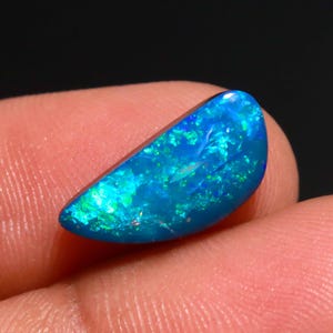 May include: A crescent-shaped loose gemstone featuring a deep blue base color with vibrant flashes of neon green and electric blue play-of-color throughout the surface. The stone has a smooth, polished finish and a curved, semi-circular silhouette.